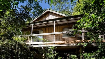 A two-story wooden cabin with a balcony, surrounded by dense forest.