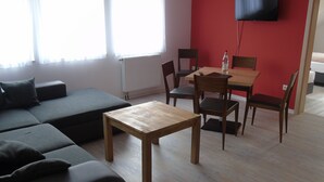 Uma sala de estar moderna com sofá, uma mesa de madeira e cadeiras. Há uma televisão fixada na parede.