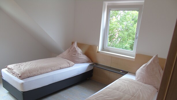 Um quarto pequeno e moderno, com duas camas de solteiro, uma janela com vista para o verde e um banco embutido.