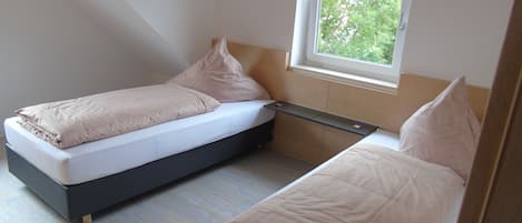 A small, modern bedroom with two single beds, a window with a view of greenery, and a built-in bench.