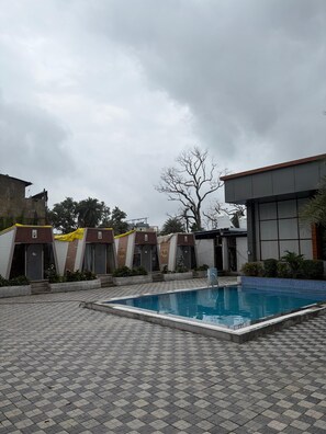 Zona de piscina con suelo de baldosas, un edificio con un ventanal grande y un árbol al fondo.