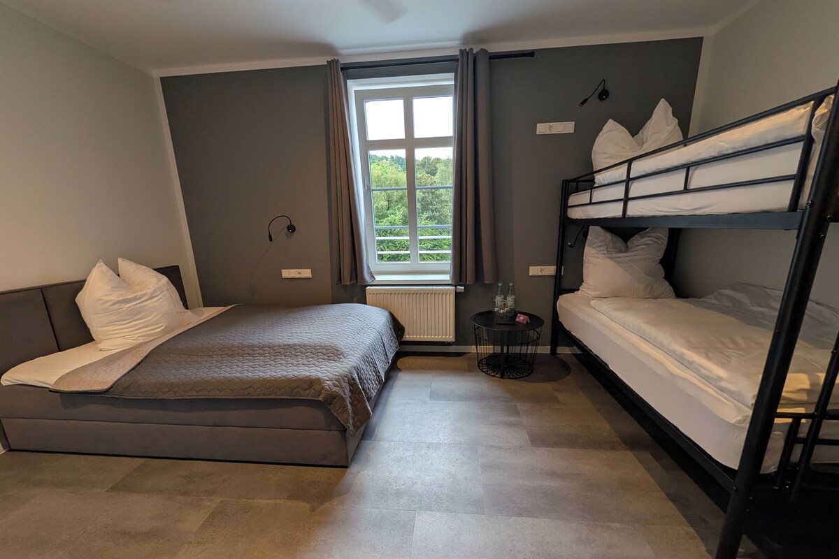 A room with a bunk bed, a single bed, a small table with bottles, a window with curtains, and a radiator.