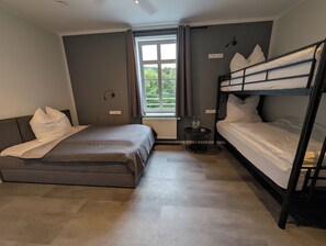 A room with a bunk bed, a single bed, a small table with bottles, a window with curtains, and a radiator.