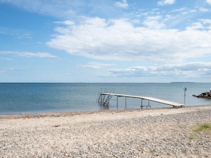 A pebbled beach with a wooden pier extending into calm waters, a bench, and a distant shoreline. - 4 Star Holiday Home in Juelsminde (Juelsminde)