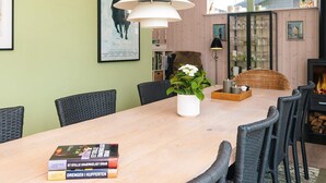 A dining area with a wooden table, wicker chairs, and a vase with flowers. There are books on the table, a fireplace, and a window with decorative items. - 4 Star Holiday Home in Juelsminde (Juelsminde)