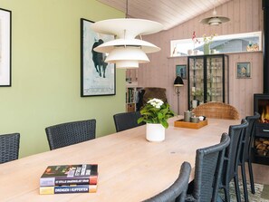 A dining area with a wooden table, wicker chairs, and a vase with flowers. There are books on the table, a fireplace, and a window with decorative items. - 4 Star Holiday Home in Juelsminde (Juelsminde)
