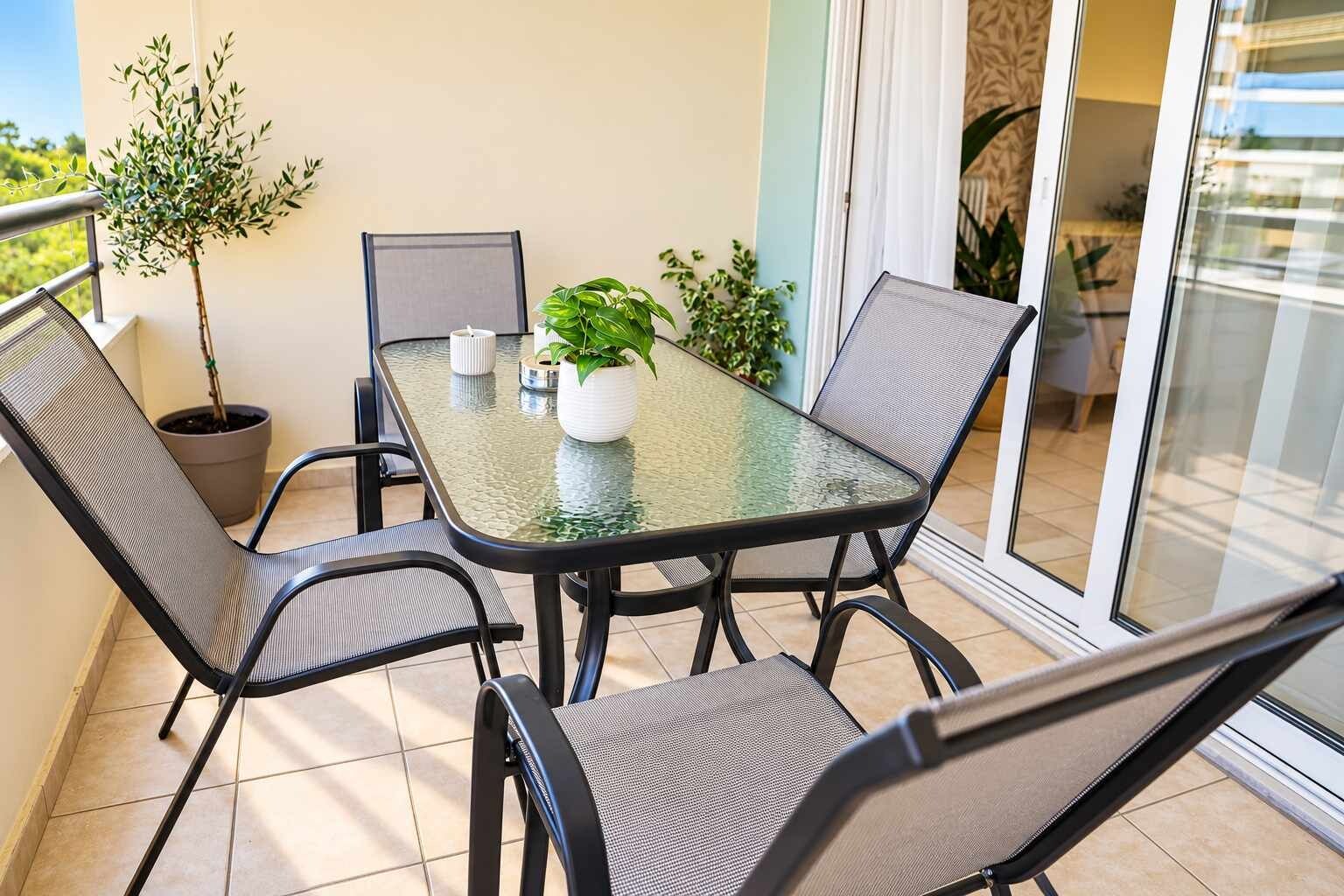 A glass-top table with four chairs, a potted plant, and a small table lamp on a balcony with a view of the sky.