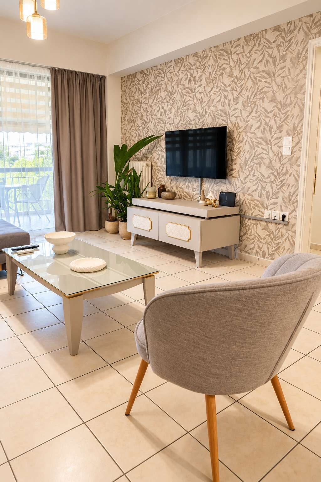 A modern living room with a glass coffee table, a grey armchair, a white console table with decorative items, a flat-screen TV mounted on the wall, and a large window with curtains.