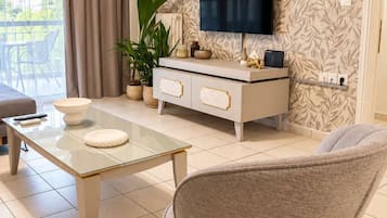 A modern living room with a glass coffee table, a grey armchair, a white console table with decorative items, a flat-screen TV mounted on the wall, and a large window with curtains.