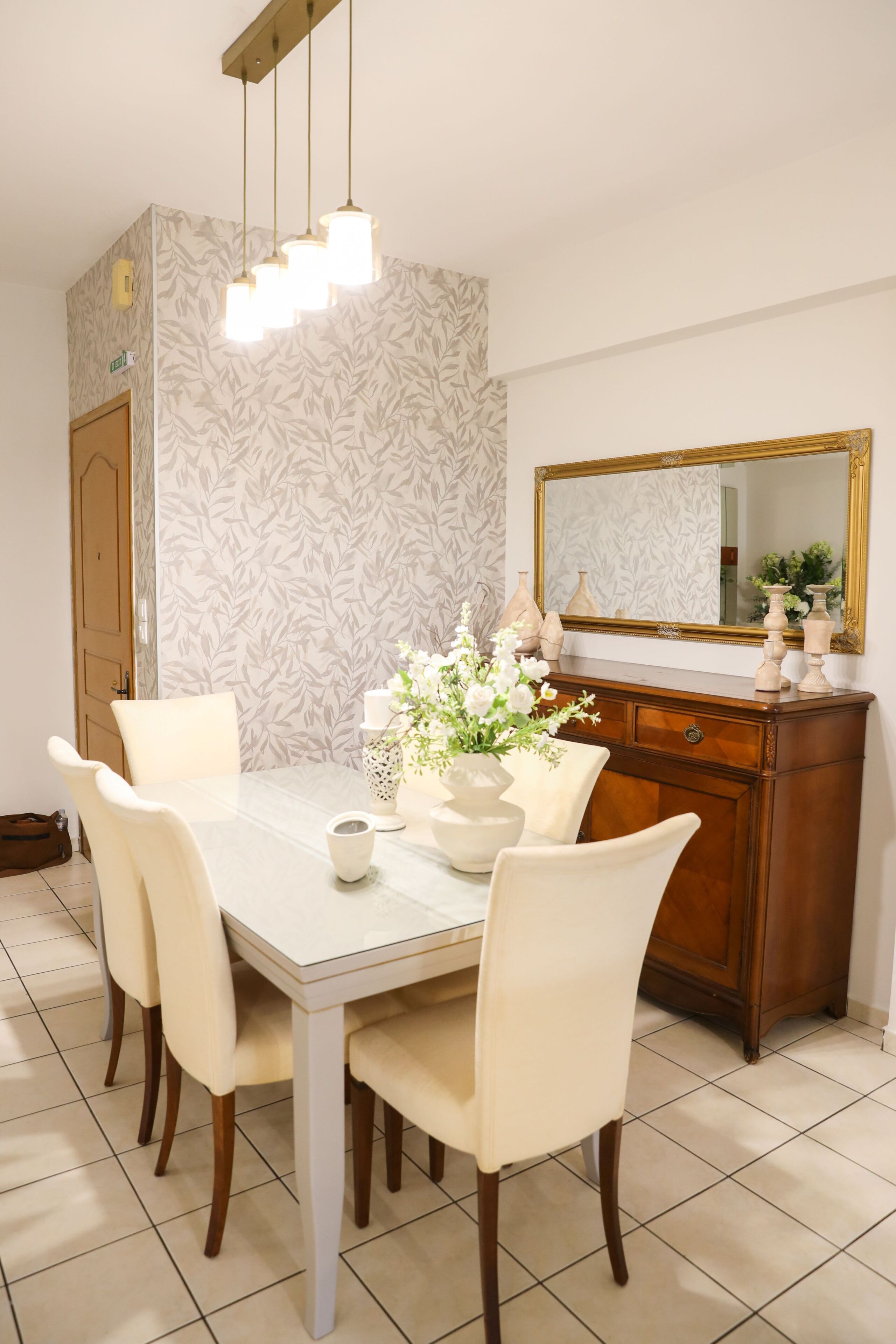 A dining area with a white table, four chairs, a vase with flowers, a mirror, and a wooden cabinet.