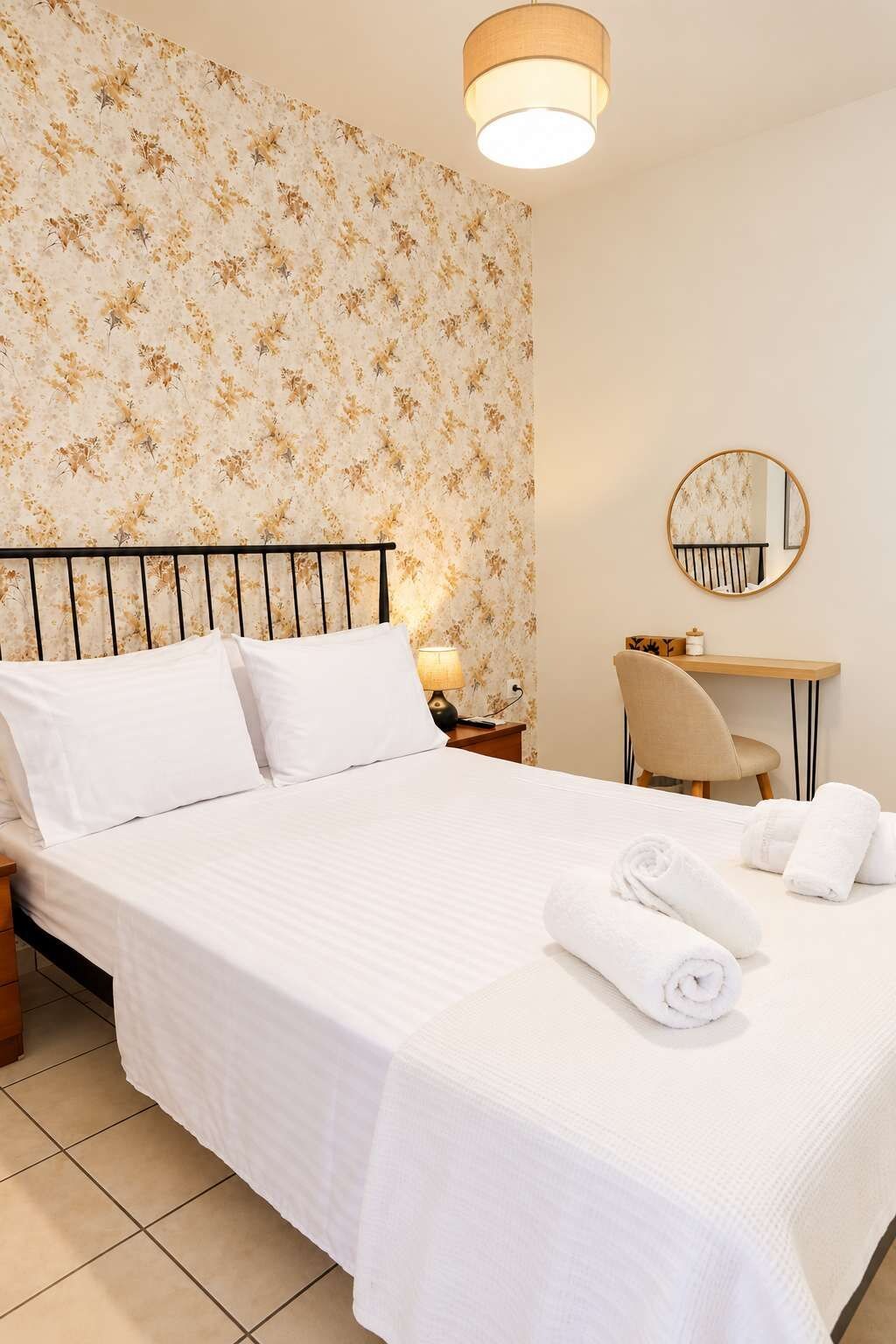 A neatly made bed with white linens, a round mirror on the wall, and a desk with a chair.