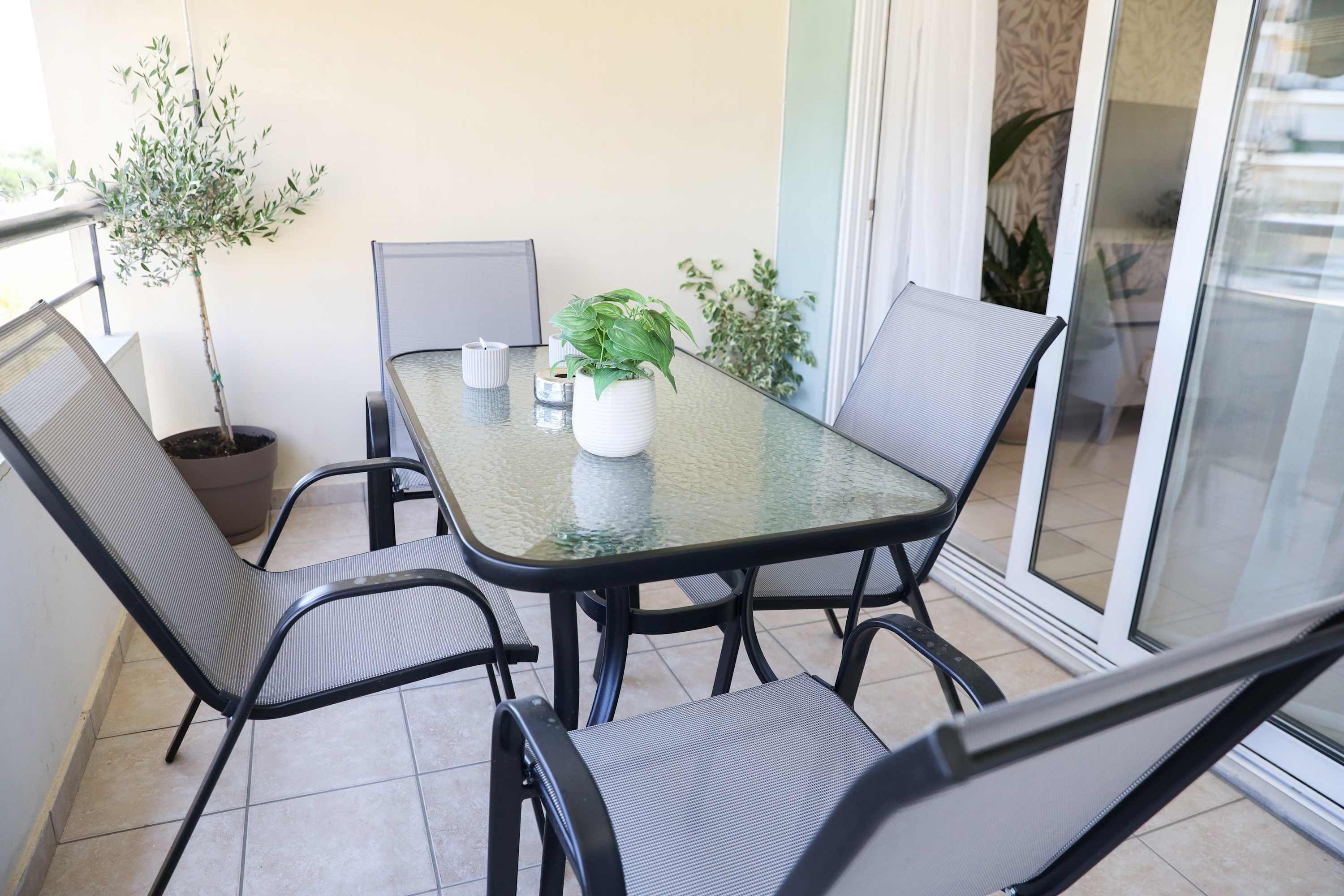 Una mesa de vidrio con cuatro sillas, una planta en maceta y una lámpara de mesa pequeña en un balcón con puerta corrediza de cristal.