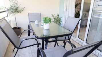 Una mesa de vidrio con cuatro sillas, una planta en maceta y una lámpara de mesa pequeña en un balcón con puerta corrediza de cristal.