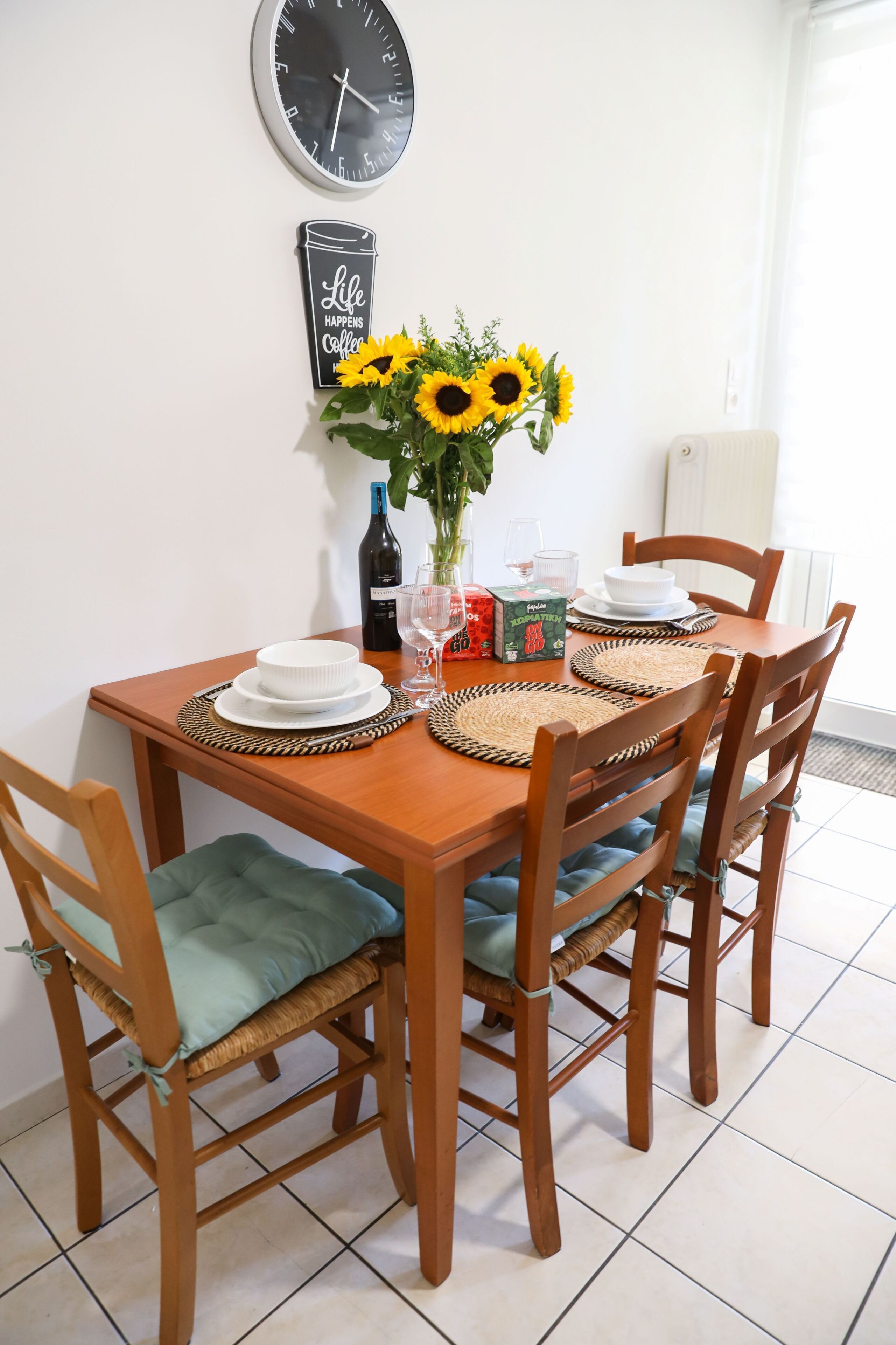 Une table à manger avec quatre chaises, un vase rempli de tournesols, une bouteille de vin et une horloge au mur.