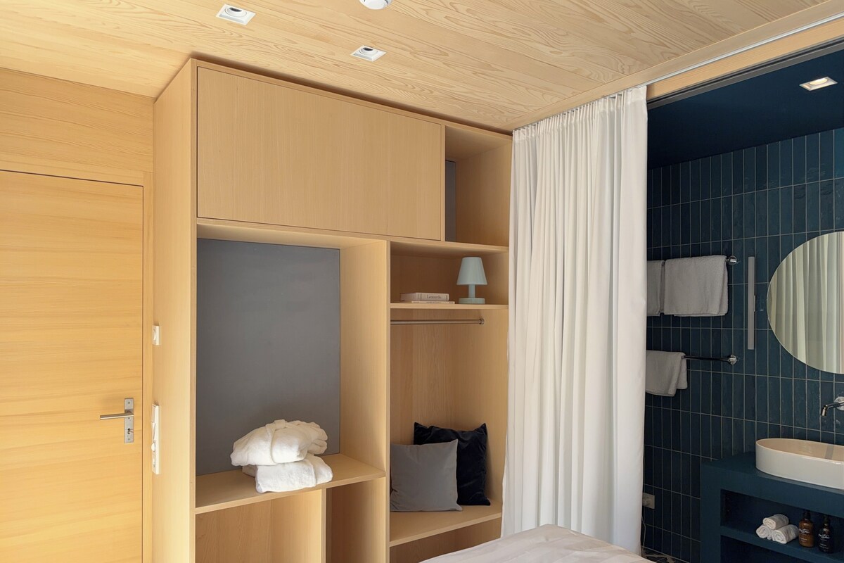 A hotel room with a wooden wardrobe, a bed with white bedding, a bedside table with a lamp, and a bathroom with a blue tiled wall and a round mirror.