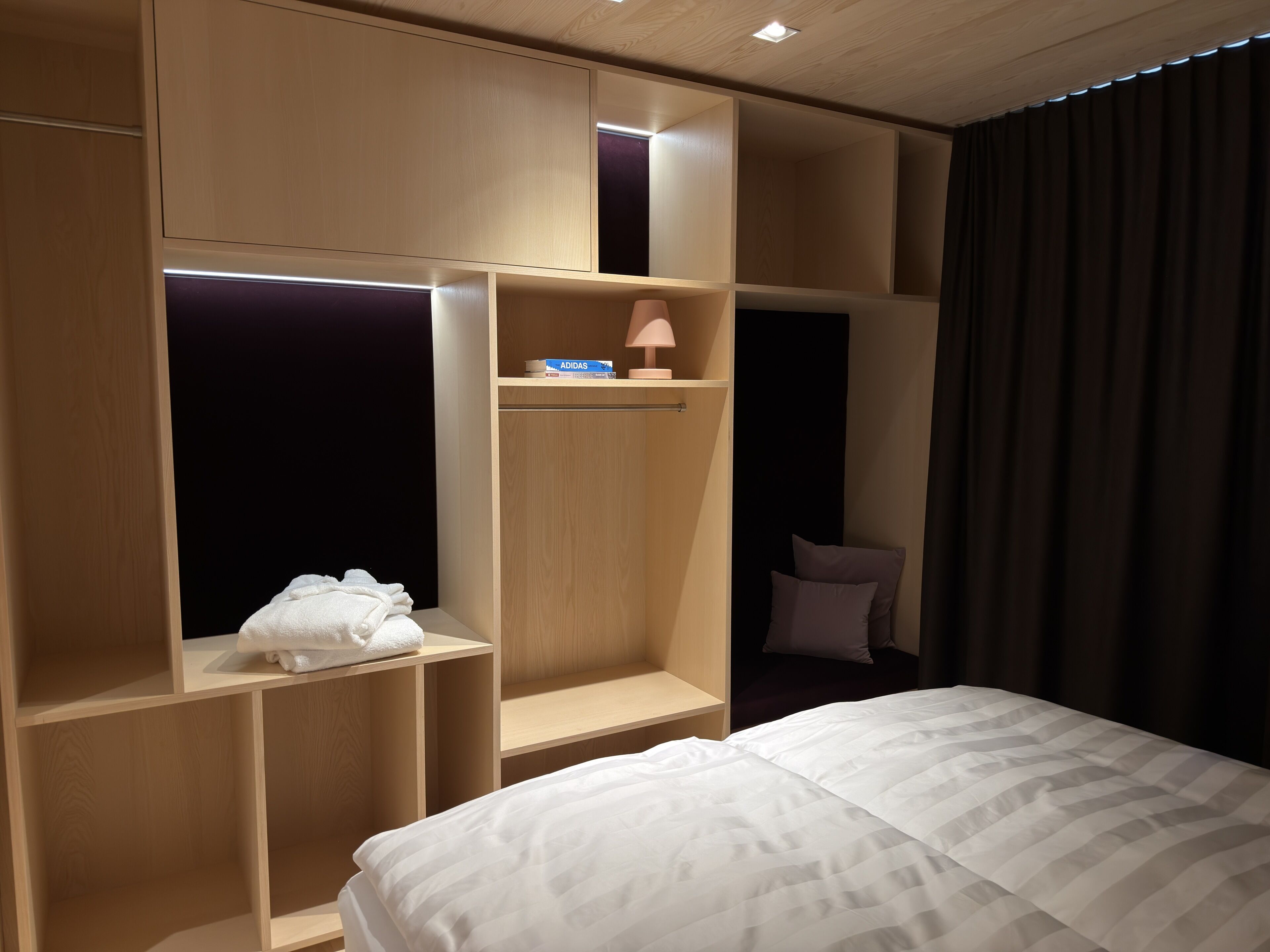 Ett hotellrum med en säng, en garderob med flera fack, en hylla med en lampa och en mörk gardin.