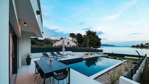 A modern outdoor pool area with a dining table and chairs, surrounded by a low wall and overlooking a body of water. - Villa Anamaria Zadarvillas (Zadar)