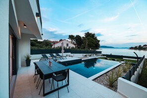 Ein moderner Poolbereich im Freien mit Esstisch und Stühlen, umgeben von einer niedrigen Mauer und Blick auf ein Gewässer.
