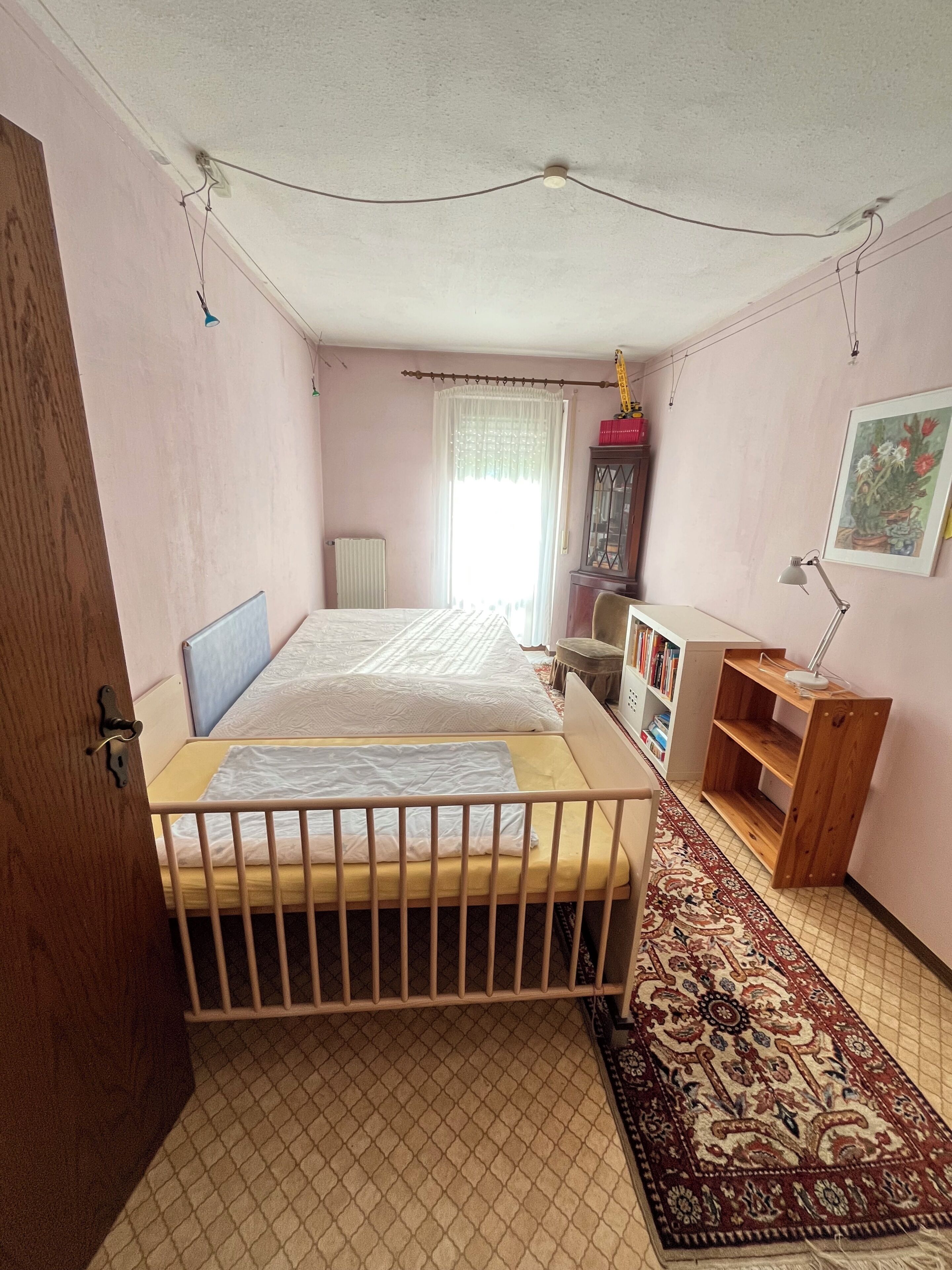 Ein Schlafzimmer mit einem Doppelbett, einem Holzschrank, einem Schreibtisch mit Lampe, einem Heizkörper und einem Fenster mit Vorhängen.