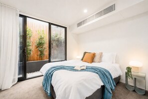 Un dormitorio con cama, una mesita de noche con lámpara, ventana con vista a una cerca de madera y plantas, y un respiradero en el techo.