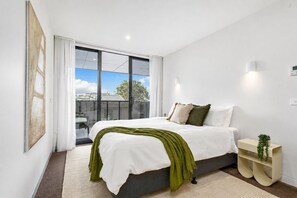Ein modernes Schlafzimmer mit einem großen Bett, Blick durch eine Glasschiebetür, einem Nachttisch mit Pflanze und einer Wandleuchte.