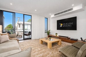 A modern living room with a large TV, a sofa, a coffee table, and a sliding glass door leading to an outdoor area. - The Richmond Grand | Near MCG + 3 Free Car Spaces (Richmond)