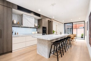 A modern kitchen with a marble countertop, wooden cabinets, and a bar area with stools. - The Richmond Grand | Near MCG + 3 Free Car Spaces (Richmond)