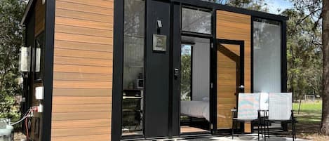 A modern, compact cabin with a wooden exterior and a glass door, situated in a wooded area.