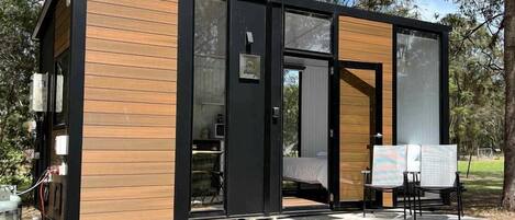 A modern, compact cabin with a wooden exterior and a glass door, situated in a wooded area.