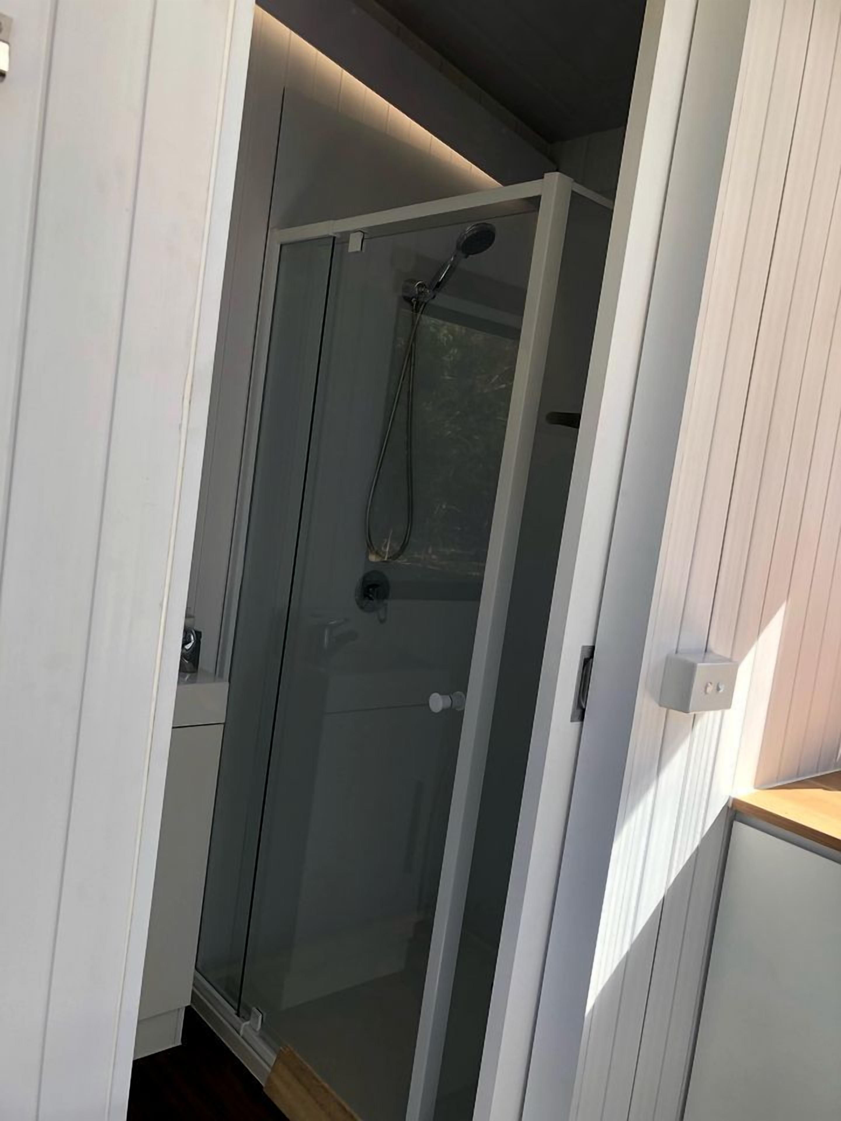 A glass-enclosed shower area with a showerhead and control panel, adjacent to a white wall with a door.
