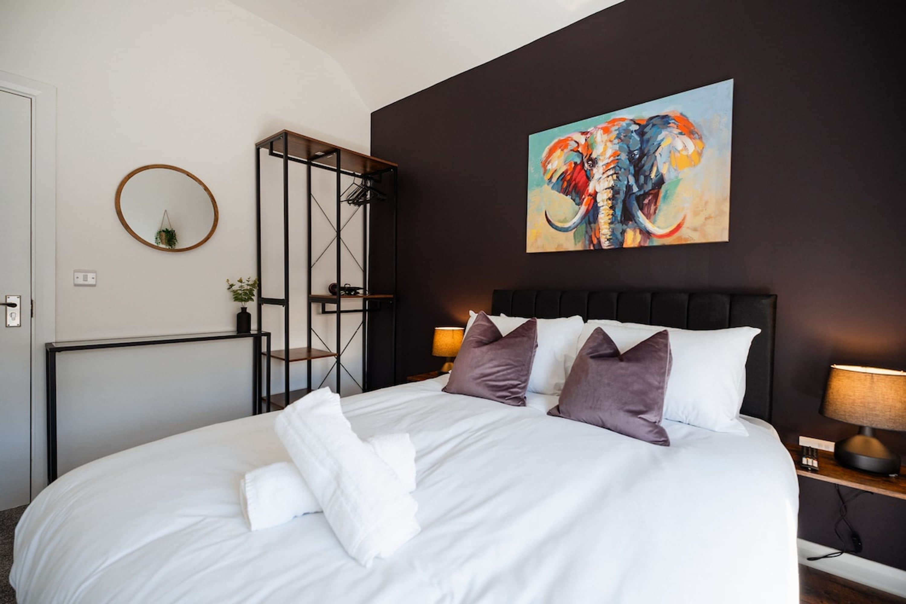 Une chambre d’hôtel avec un grand lit, une tête de lit, un tableau, un miroir rond et une petite plante.