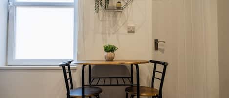 Un petit coin repas avec une table et deux chaises, une plante en pot posée sur la table et une fenêtre avec des stores.