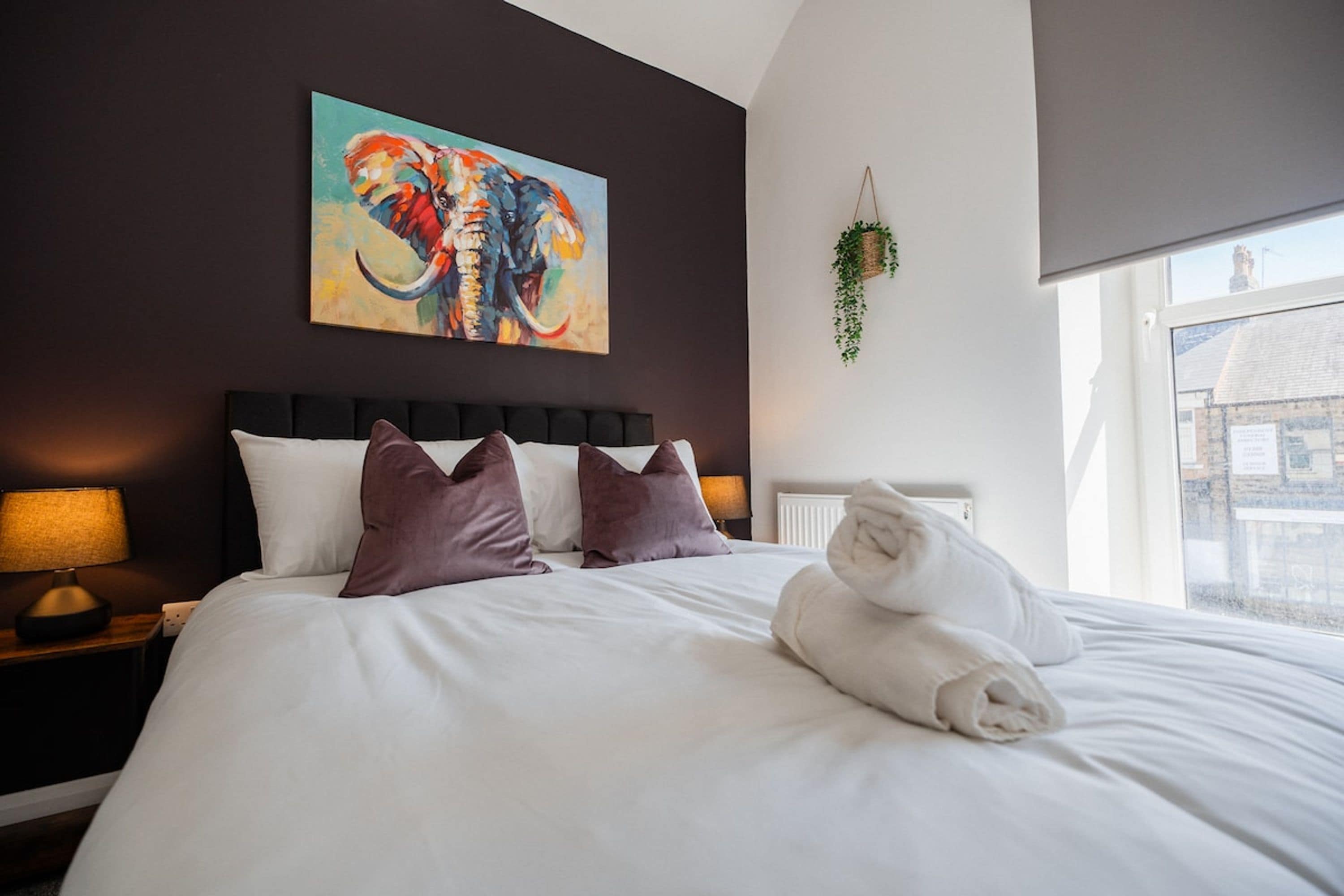 Ein Hotelzimmer mit einem großen Bett, einem Elefantenbild, einer hängenden Pflanze und einem Fenster mit Blick auf Gebäude.