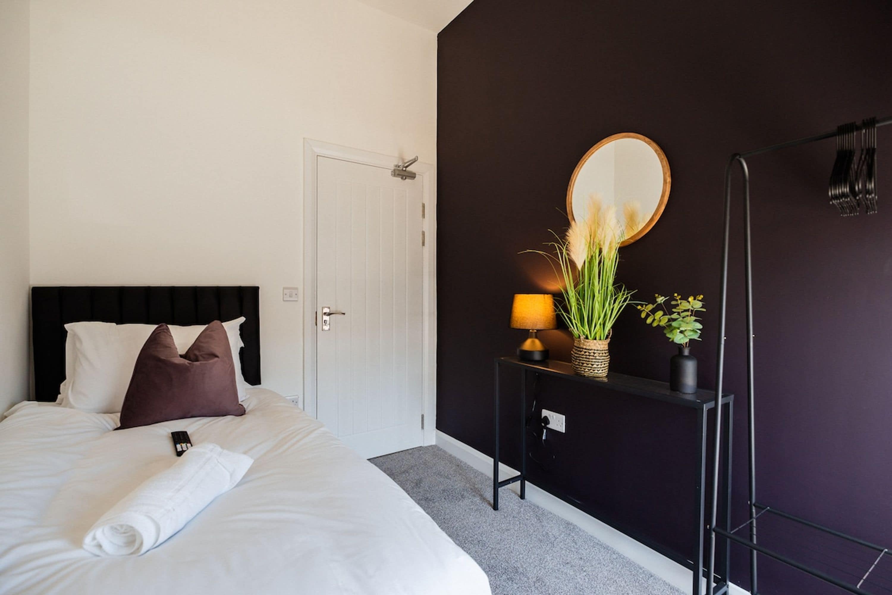 A hotel room with a bed, a bedside table with a lamp, a mirror, and a wall-mounted rack.