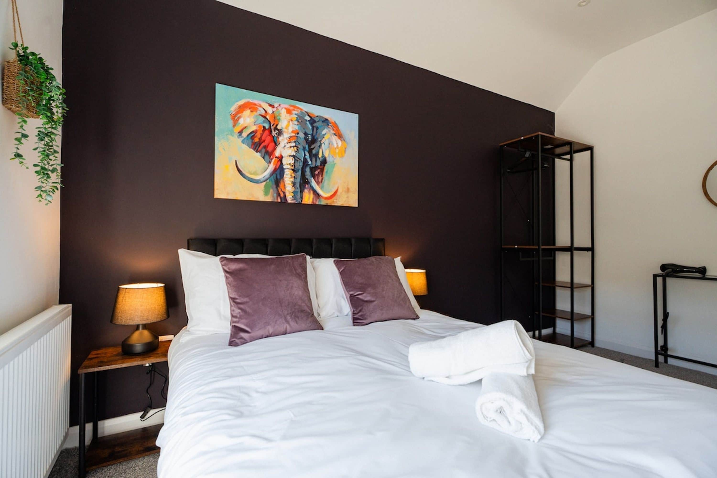Une chambre avec un grand lit, deux lampes de chevet, une œuvre d’art représentant un éléphant accrochée au mur, et une plante sur le côté gauche.