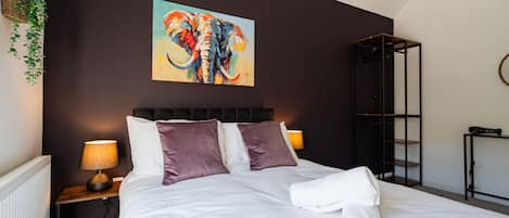 A bedroom with a large bed, two bedside lamps, a wall-mounted artwork of an elephant, and a plant on the left.