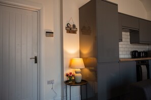Een keuken met een witte deur, een bord aan de muur, een tafel met een lamp en bloemen, en een aanrecht met keukenapparatuur.