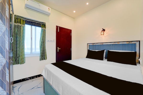 A hotel room with a bed, a window with curtains, an air conditioning unit, and a wall-mounted light fixture.