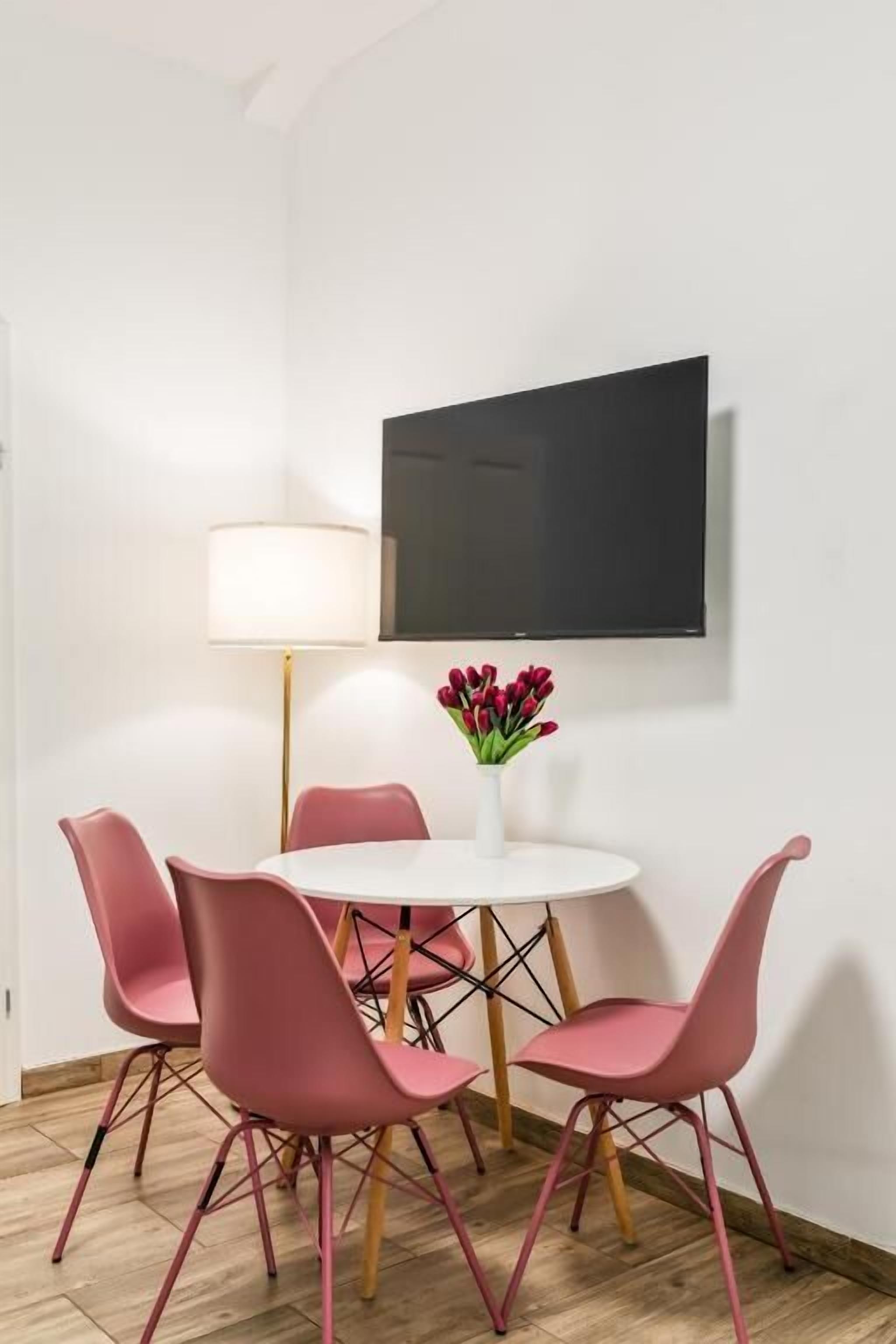 Una habitación con una mesa redonda, cuatro sillas rosas, un florero blanco con flores, una lámpara de pie y un televisor de pantalla plana montado en la pared.