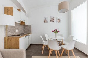 Cocina moderna con gabinetes blancos, encimera de madera y un lavamanos. Hay un comedor con una mesa y sillas blancas, y un jarrón con flores.