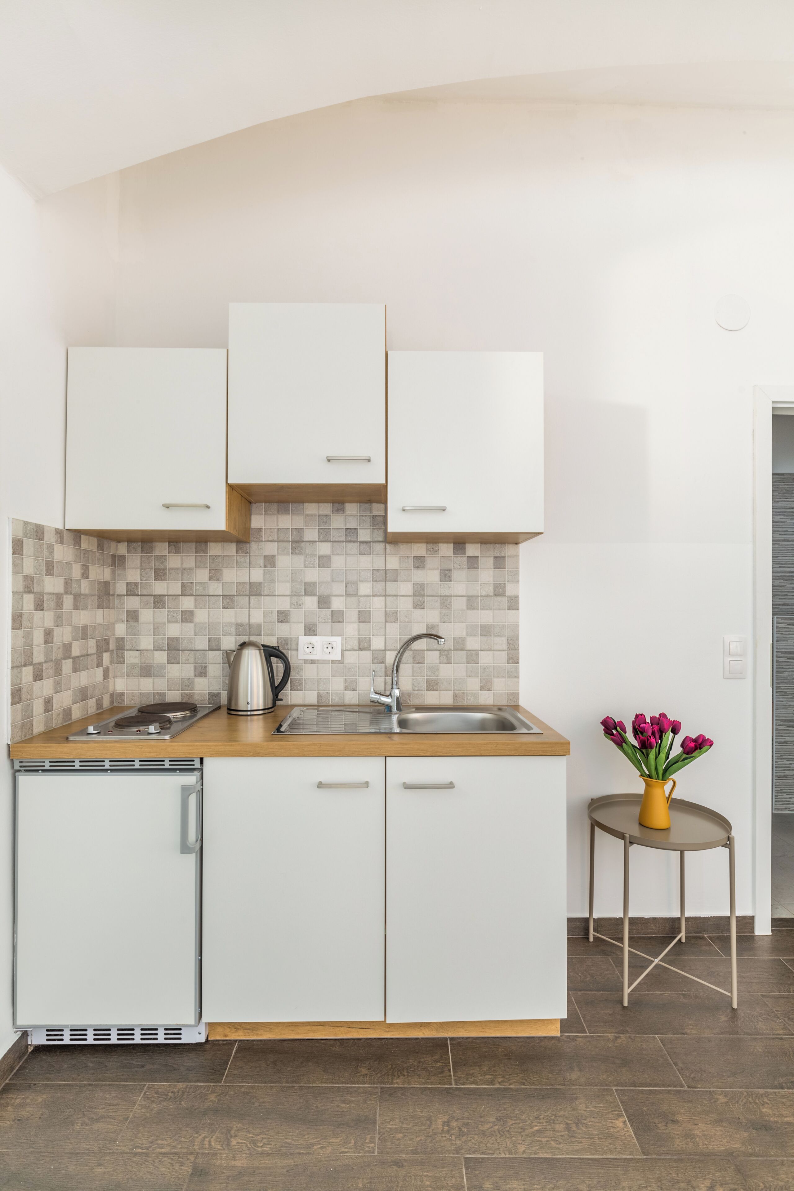 A modern kitchen with white cabinets, a tiled backsplash, a stainless steel kettle, and a small round table with a vase of flowers.