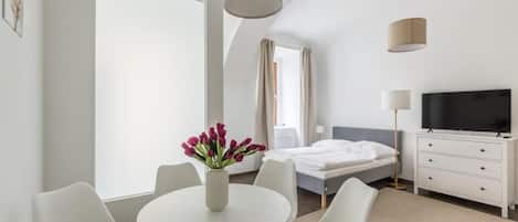 A modern hotel room with a bed, a TV, a desk with chairs, and a vase of flowers.