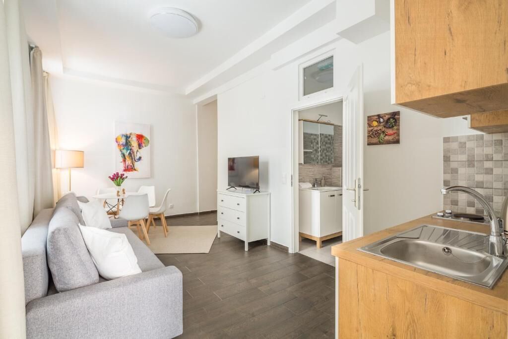 Ein modernes Wohnzimmer mit grauem Sofa, ein Essbereich mit weißem Tisch und Stühlen, eine Küche mit Holz Arbeitsplatte und Edelstahlspüle sowie eine weiße Wand mit einem farbenfrohen abstrakten Gemälde.
