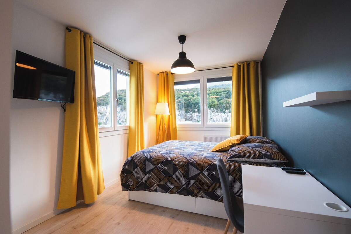 A bedroom with a bed, a TV mounted on the wall, a window with curtains, and a lamp.