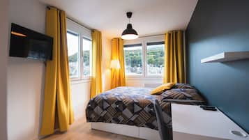 A bedroom with a bed, a TV mounted on the wall, a window with curtains, and a lamp.