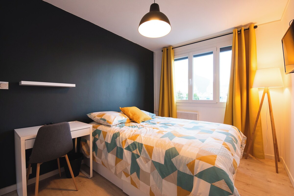 A bedroom with a bed, a desk, a chair, a lamp, and a window with curtains.