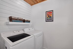 A laundry room with two white washers and dryers, wooden shelves above, and a Florida travel poster on the wall. - Tides & Turtles · Canal Dock · Waterfront (Holmes Beach)