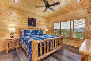 A wooden cabin bedroom with a bed, two bedside lamps, a ceiling fan, a window with blinds, and a deer antler artwork on the wall. - Jump on Inn to this luxurious new pool cabin. (Sevierville)