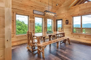 A wooden cabin interior with a large dining table, multiple chairs, and large windows offering views of a forested landscape. - Jump on Inn to this luxurious new pool cabin. (Sevierville)