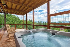 A wooden deck with a hot tub, wooden chairs, and a view of a forested landscape. - Jump on Inn to this luxurious new pool cabin. (Sevierville)
